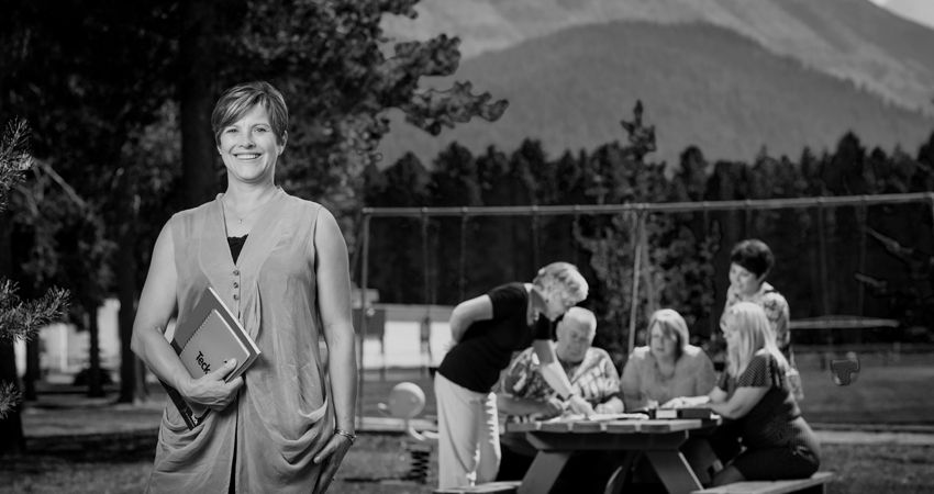 Smiling person with a tablet stands in front of a group sitting at a picnic table in a park.